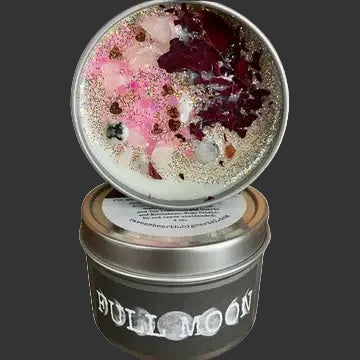 A metal tin candle shown open with the lid resting on top, revealing a white wax base with embedded pink and burgundy dried flower petals and glitter. The lower tin has a dark charcoal label reading 'BULL MOON'.