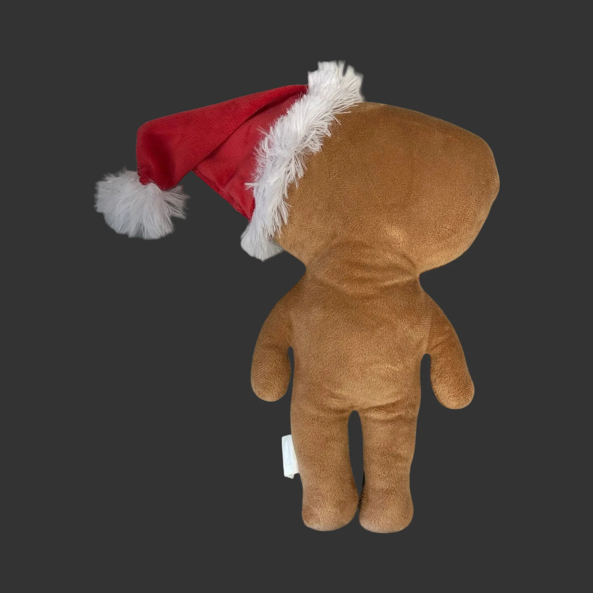 A gingerbread brown Christmas plush with a large head and simple limbs, wearing a red Santa hat with white faux-fur trim and pom-pom, photographed on a clean black background.