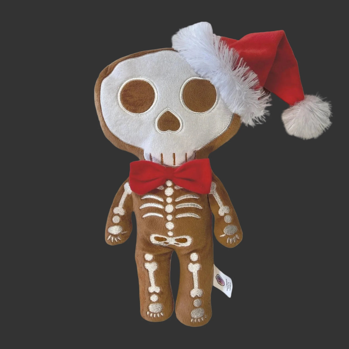 A plush toy shaped like a gingerbread skeleton wearing a red Santa hat with white trim and a red bow tie, photographed on a plain white background.