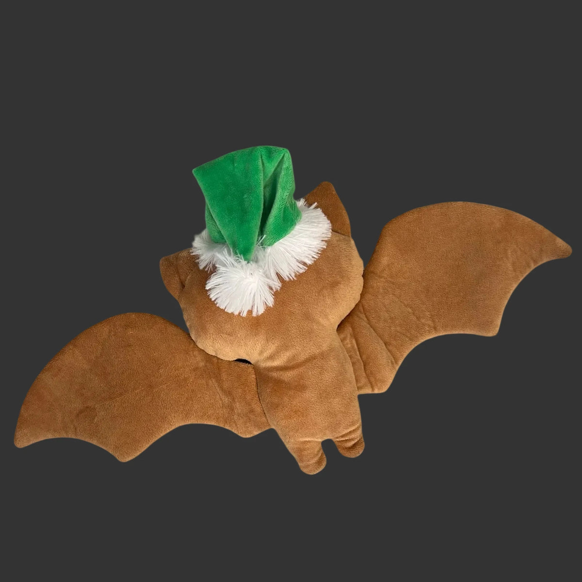 A brown plush skeleton bat soft ginger dead creepmus decoration with wings spread, wearing a green Santa hat with white faux fur trim, photographed on a plain white background.