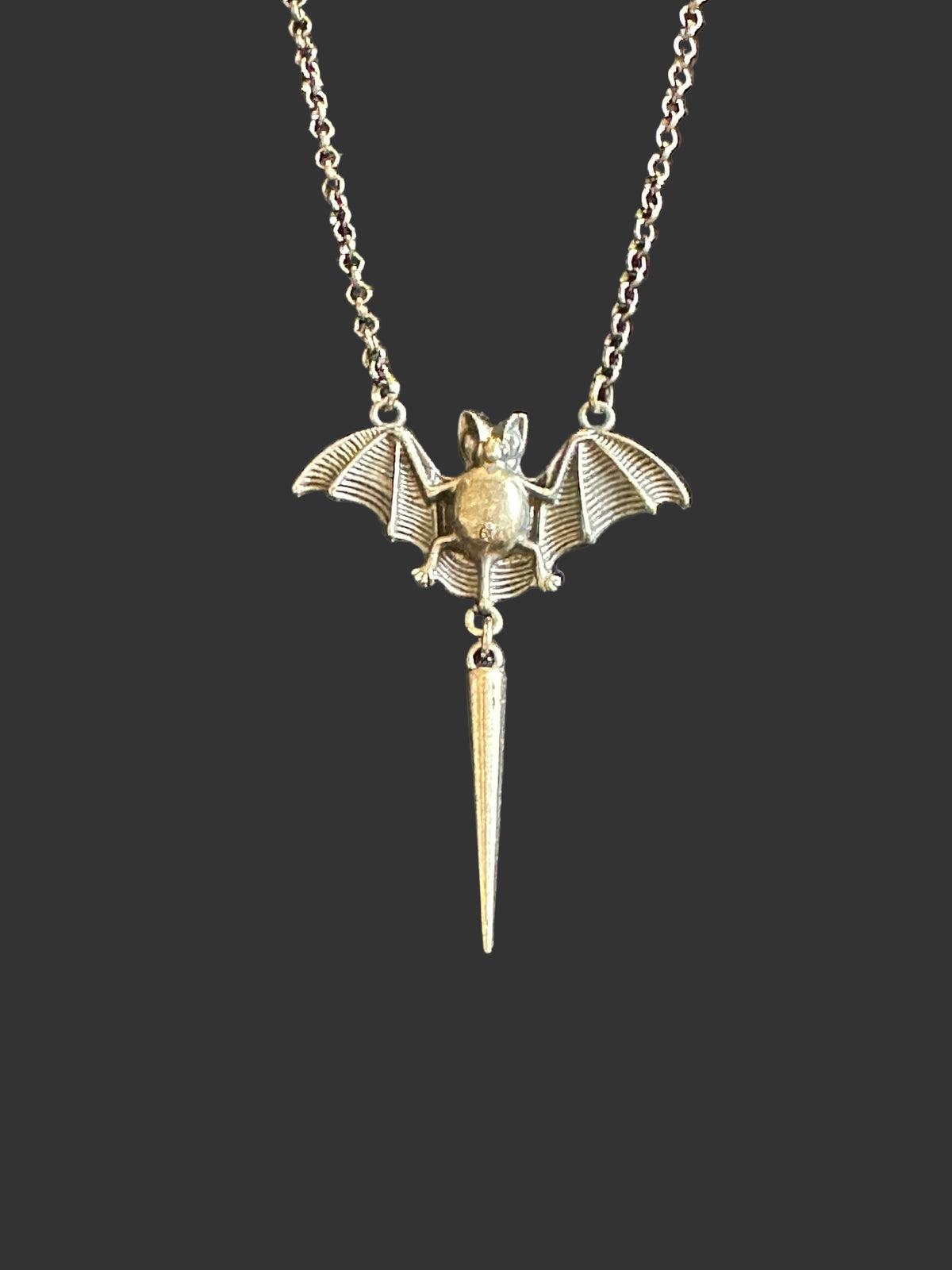 A silver-toned bat pendant necklace with spread wings and a dangling pointed bat-shaped charm, displayed on a textured dark gray fabric background.