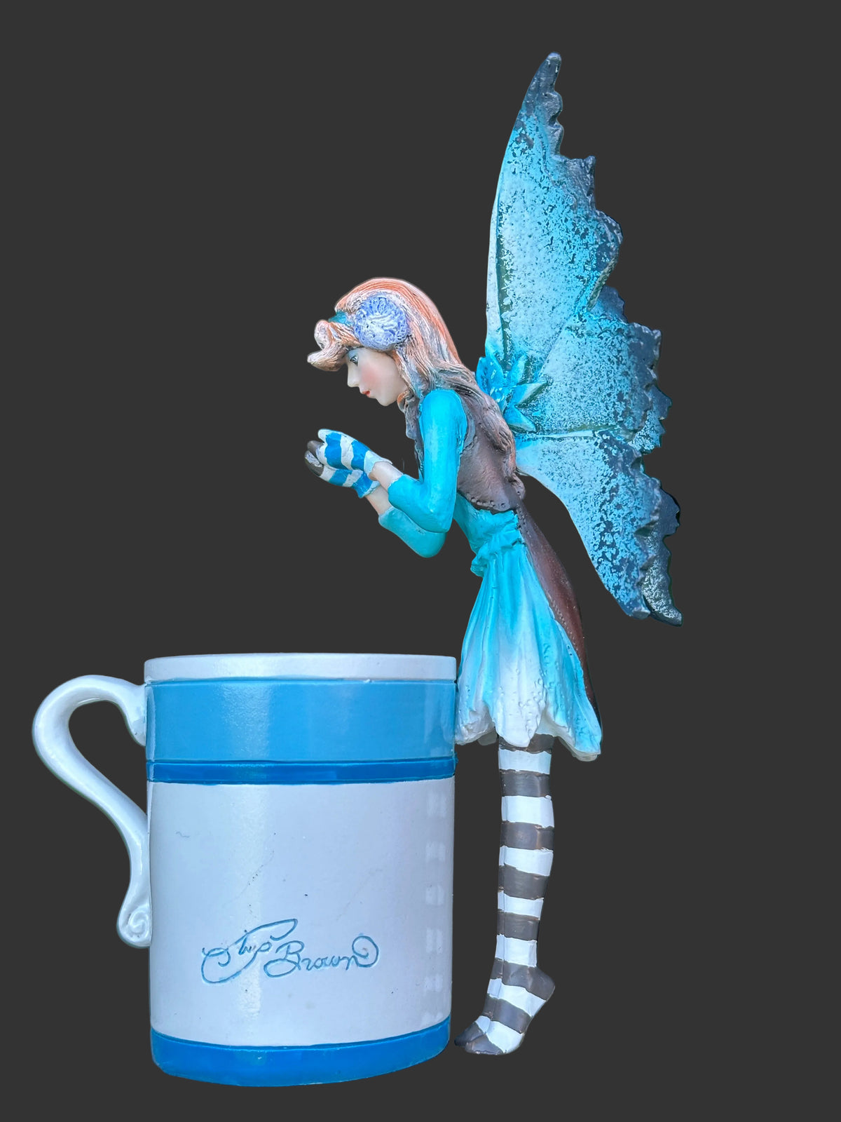 A small Amy Brown fairy figurine in a blue and white dress with striped legs stands beside a white ceramic mug with blue bands and a visible 'Amy Brown signature' logo, set against a blurred green garden background.