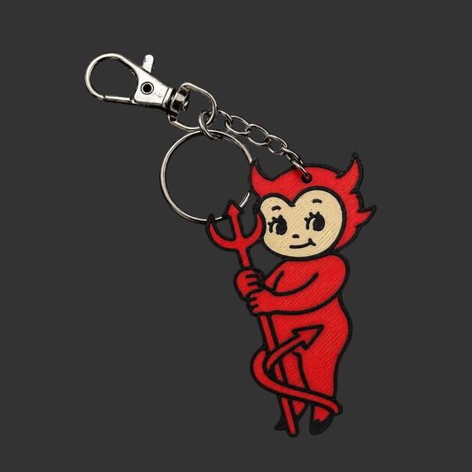 A devil-themed 3D printed keychain featuring a red vinyl charm of a cartoon kewpie style devil holding a trident, attached to a silver metal clasp and keyring, displayed on a round black slate coaster.