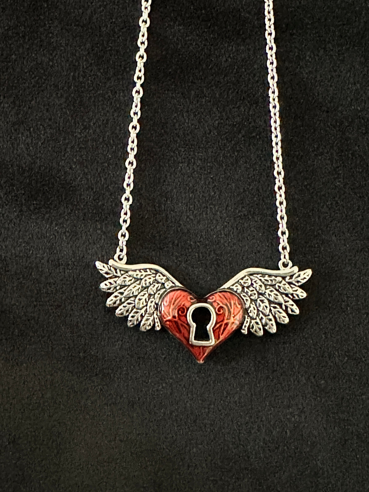 A silver-toned winged heart pendant necklace with a red enamel heart and a keyhole cutout in the center, displayed on a black textured background.