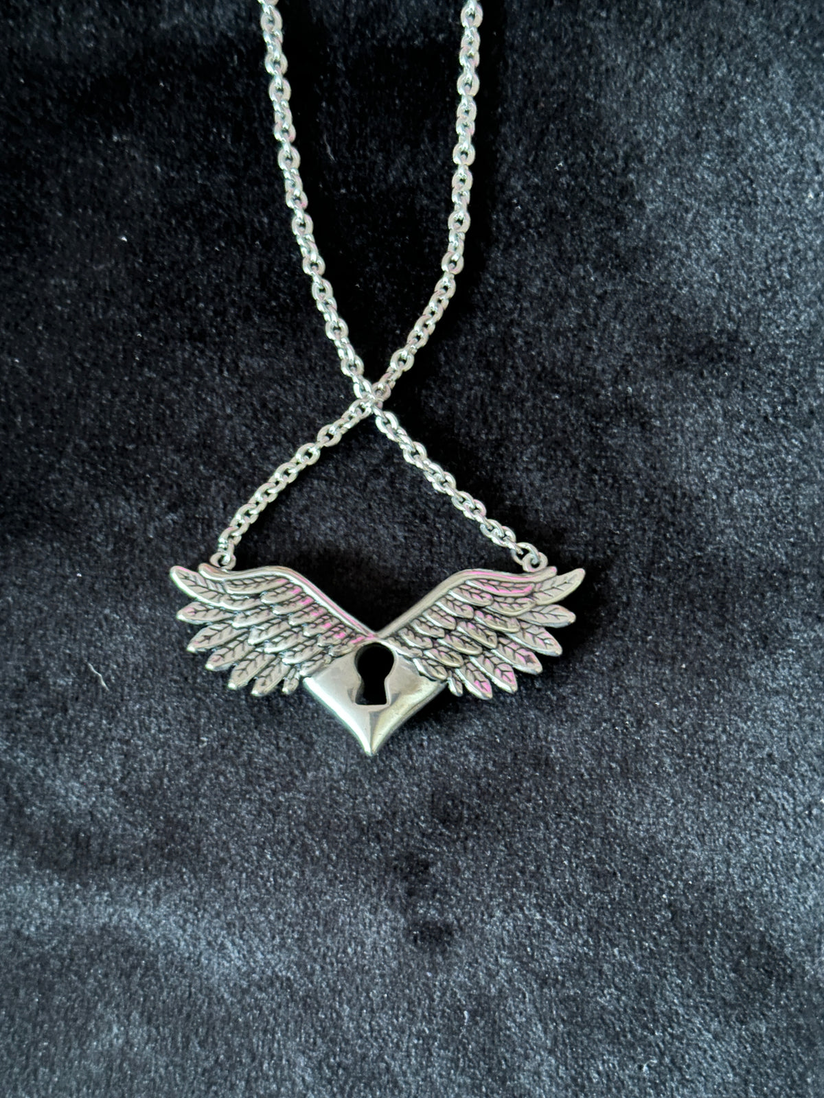 A silver-toned winged back side of pendant necklace with a keyhole detail, suspended from a matching chain against a dark textured fabric background.
