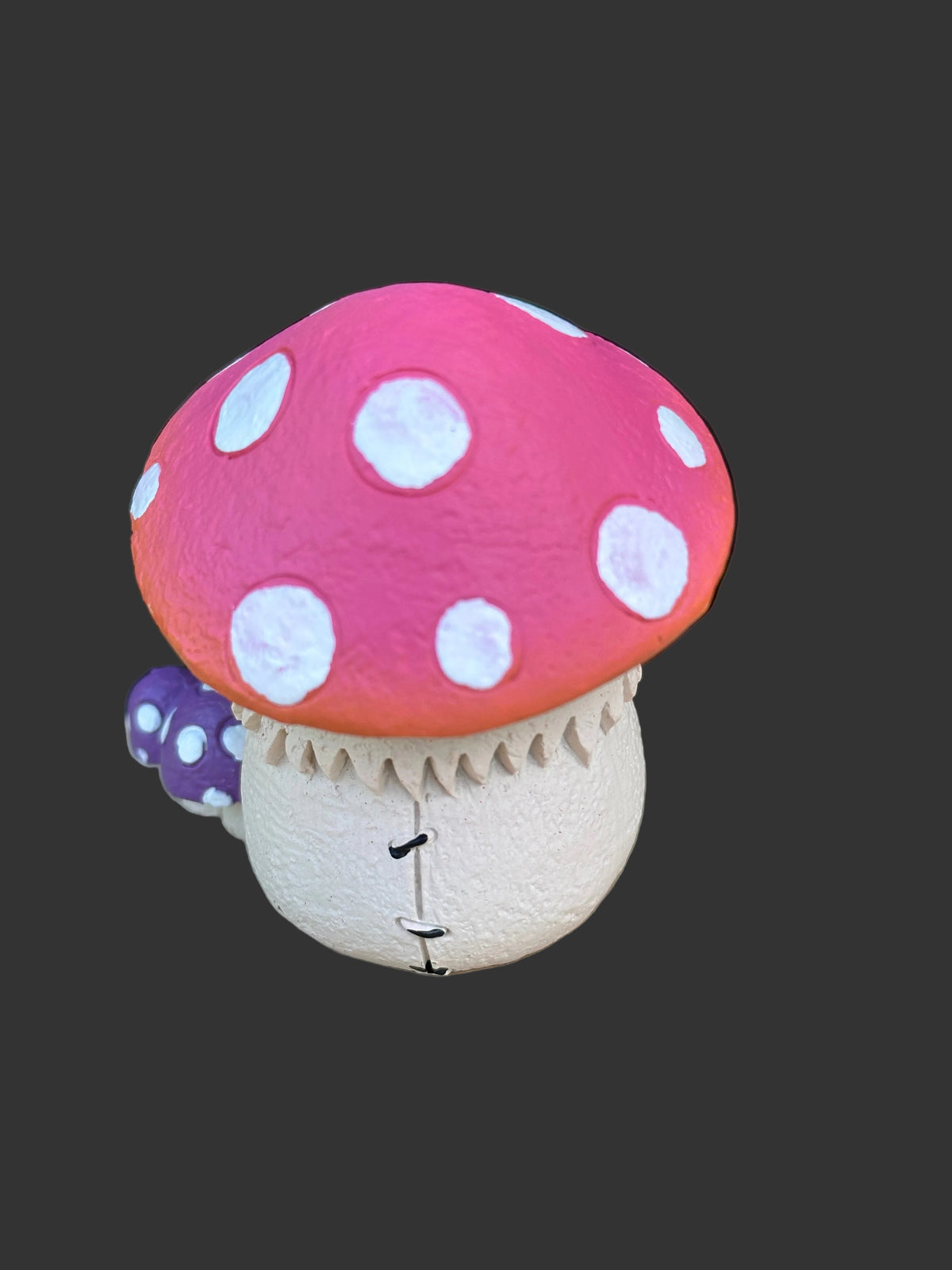 A mushroom-shaped Furrybones collection figurines with a bright pink cap, white polka dots, a cream-colored stem, and a small purple accent on the side, shown against a dark gray background.