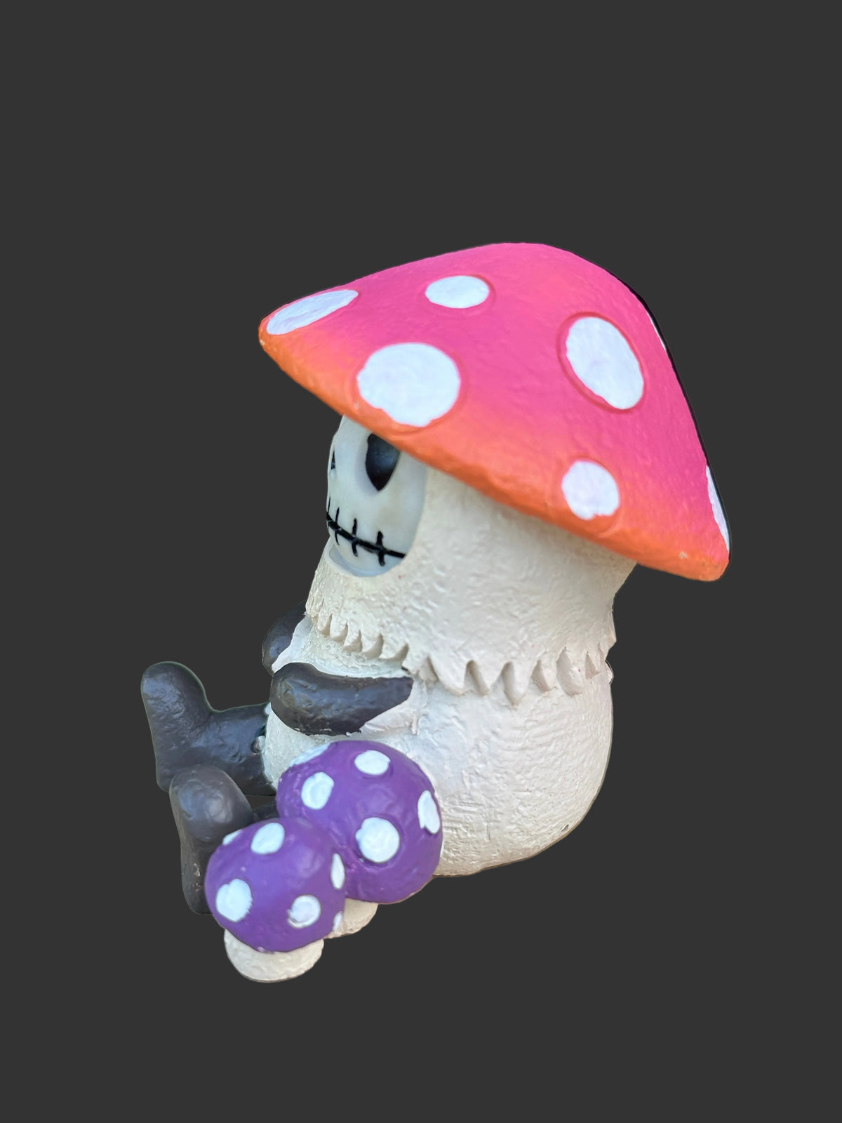 A small Furrybones figurine of a stitched skeleton character sitting inside a red-orange mushroom cap with white spots, holding two purple mushrooms with white spots, set against a solid dark gray background.
