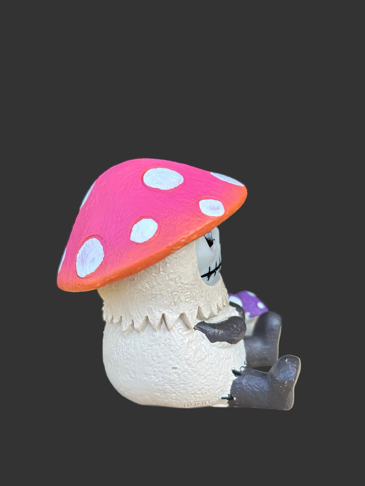 A small Furrybones mushroom-shaped figurine with a pink cap, white polka dots, an off-white textured body, and dark gray limbs, shown against a solid dark gray background.