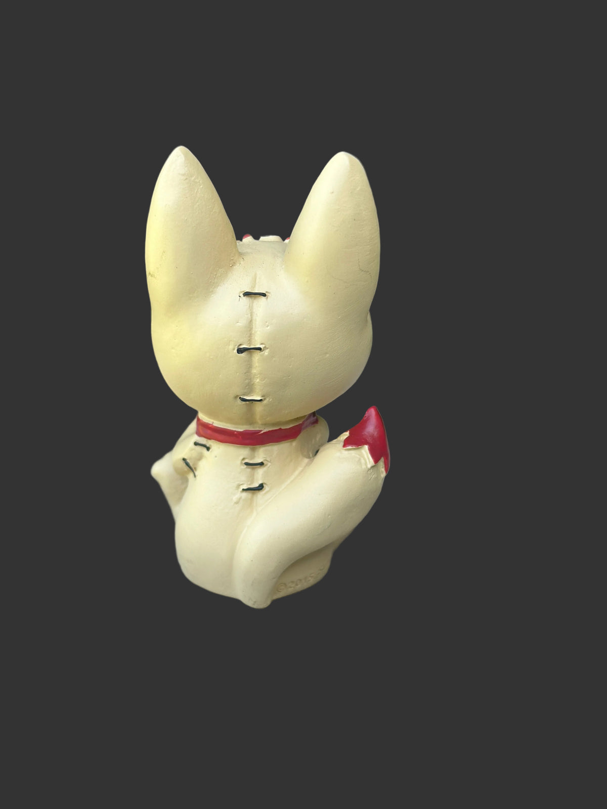 A small beige kitsune fox figurine shown from the back, sitting on a weathered gray wooden surface with a blurred green garden background. The furrybones figurine has black stitched details, a red collar, and a red paw pointing outward.