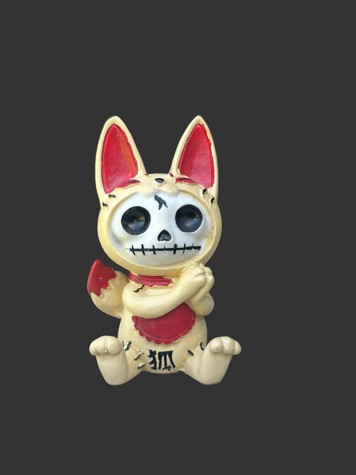 A small decorative furrybones igurine of a stylized Kitsune fox character with large ears, a skeletal face, and red painted accents, sitting on a weathered wooden surface with blurred green foliage in the background.
