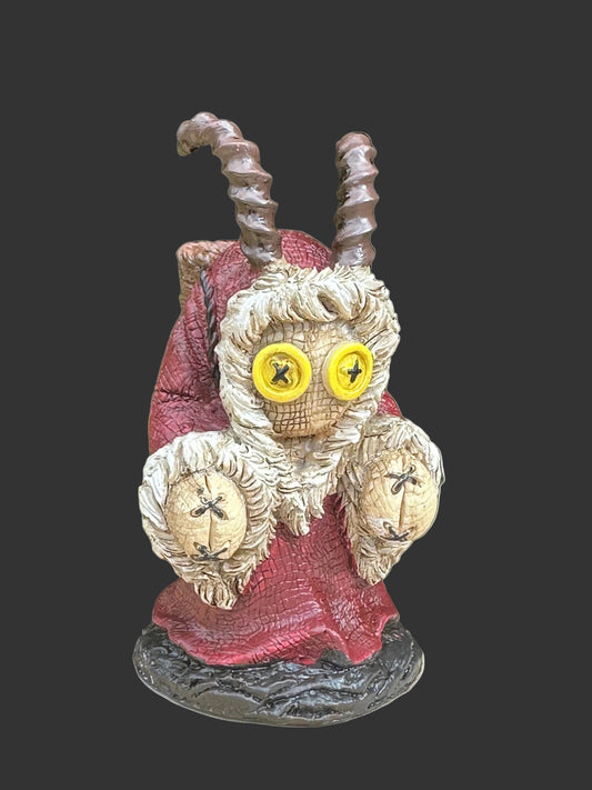 A whimsical goat-like figurine known as Krampus or statue with large yellow button eyes, textured fur, twisted horns, and a red robe, displayed outdoors on a wooden surface with green foliage in the background. Pinhead Monster