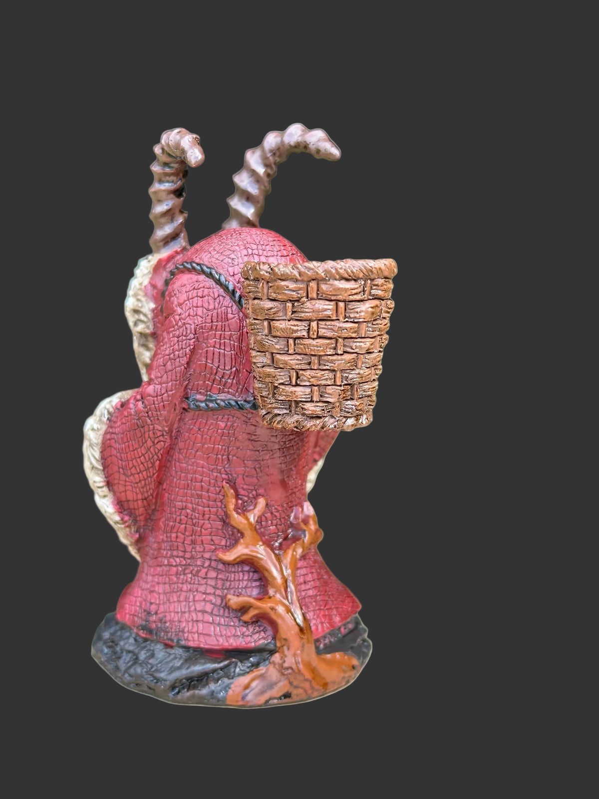 A small collectible Pinhead monster figurine of a hooded figure known as Krampus with a basket on its head, standing on a tree stump with a blurred green garden background.