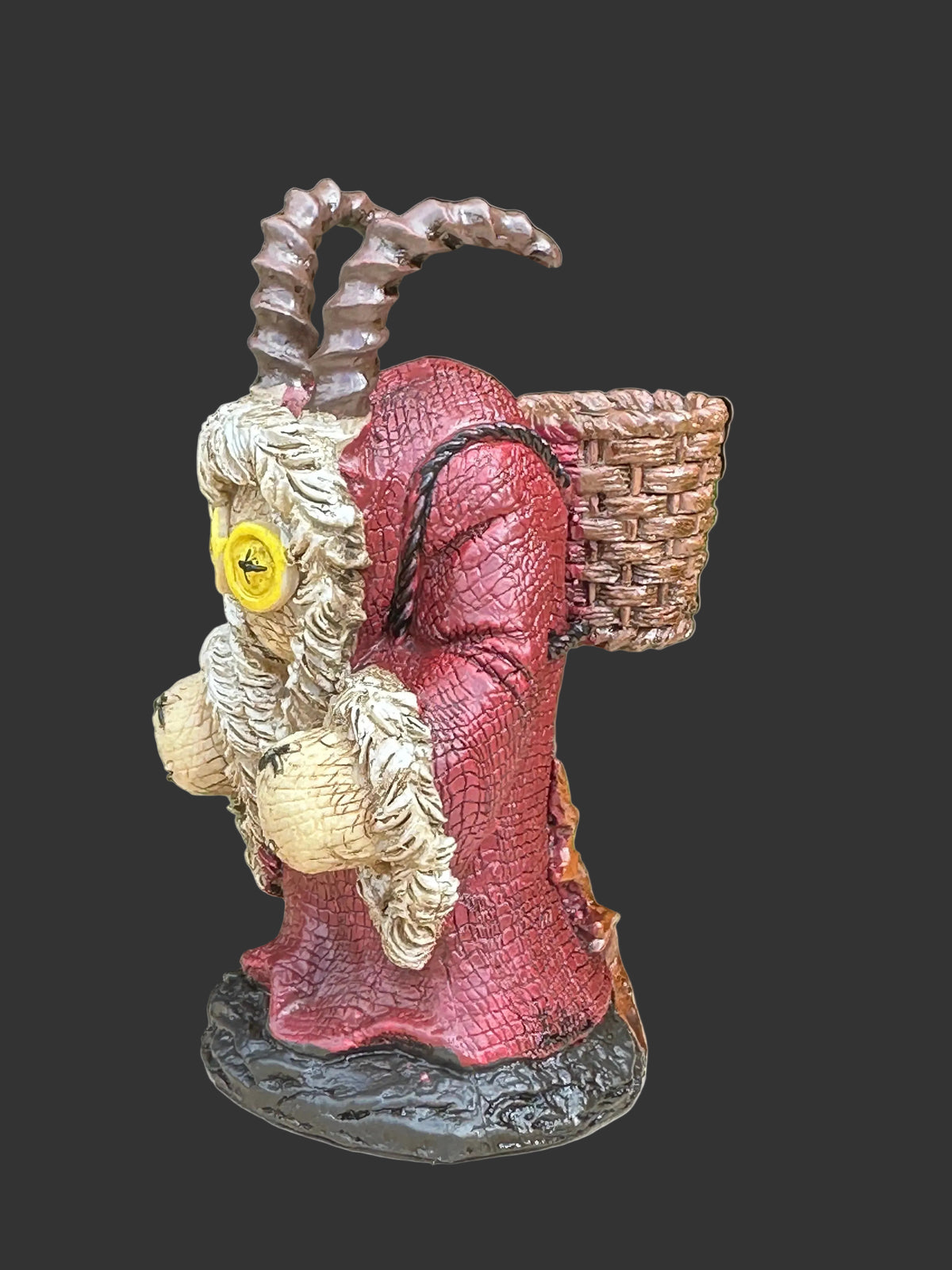 A creepy goat-like  figurine. known as Krampus with curved horns, a red robe, white beard, and a woven basket on its back, displayed during the holidays or year around on a tree stump with green foliage in the background.