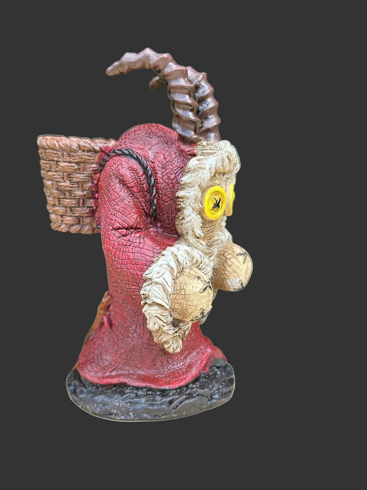A small Pinhead Monster collectible figurine of a horned creature known as Krampus wearing a red robe and carrying a basket, standing on a tree stump with a blurred green outdoor background.