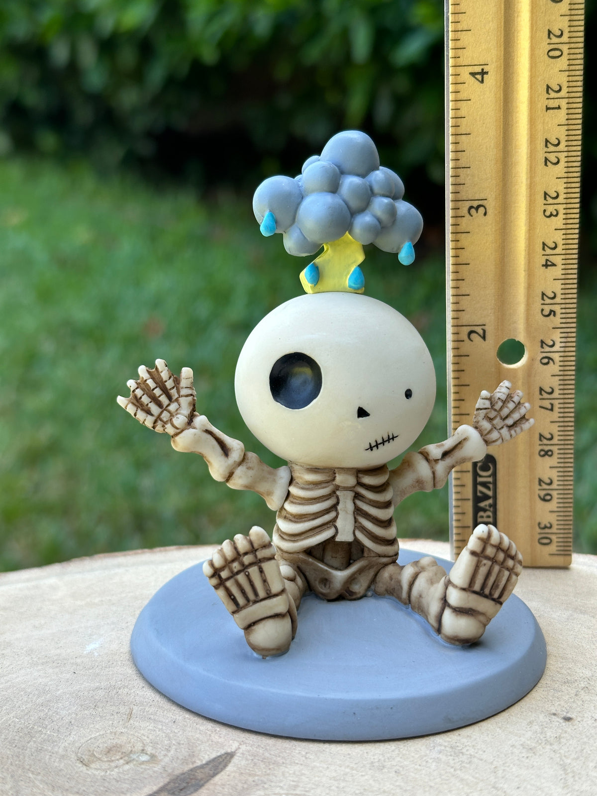 A small unlucky Luckyz skeleton Eustis figurine sitting on a round blue base, with a gray cloud on its head and a wooden ruler standing beside it for scale, photographed outdoors on a stone surface with blurred greenery in the background.