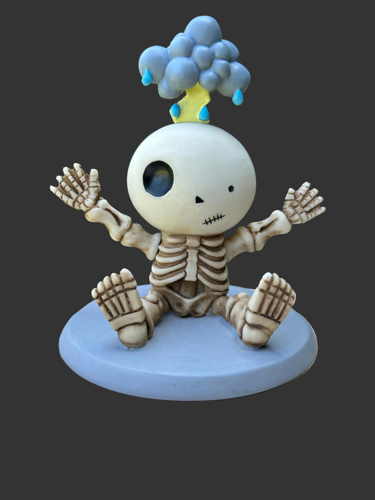 A cute Luckyz collectible skeleton figurine sitting on a round light blue base, with its arms raised and a blue cloud with yellow strike of lightening on its head, set against a dark gray background.