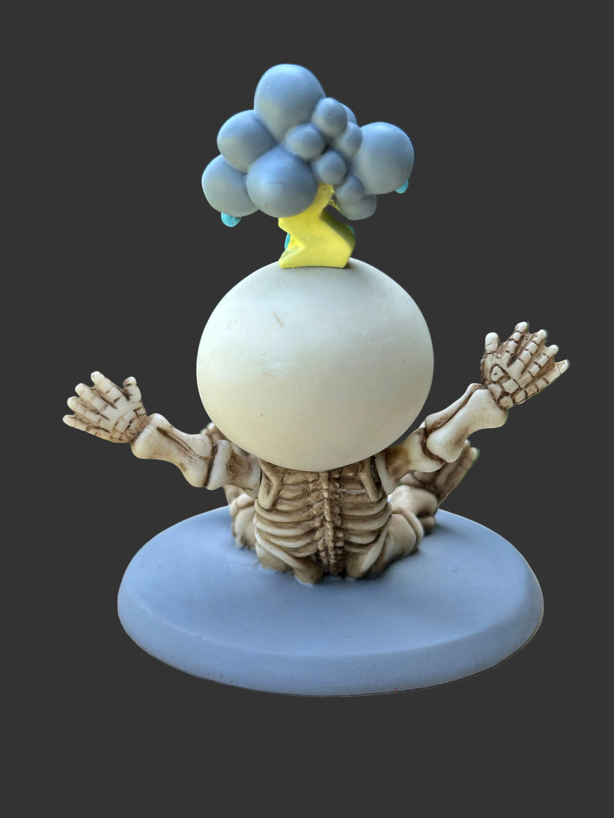 A whimsical figurine of a skeleton sitting on a round blue base, with a large white egg-shaped head, blue clouds, and a small yellow tree on top, set against a dark gray background.