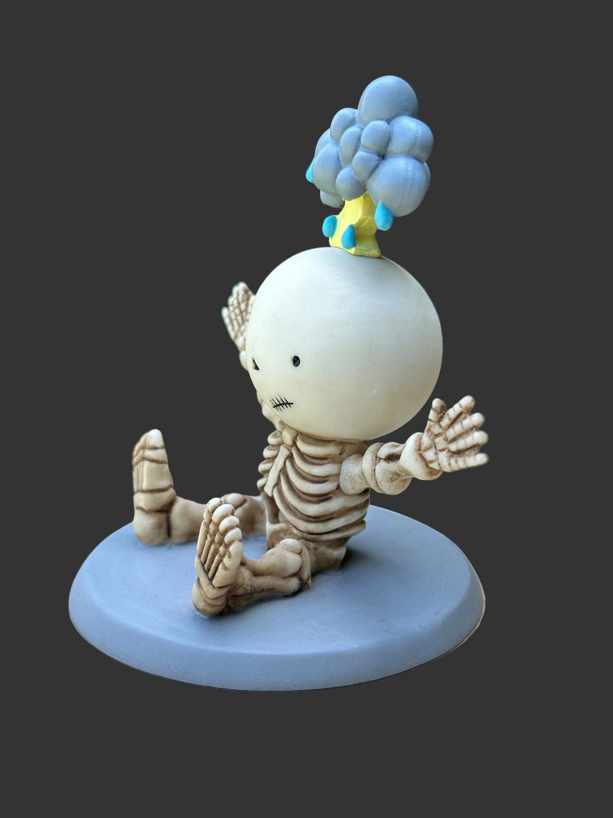 A small figurine of a seated skeleton-like character on a round pale blue base, with a round white head, beige bones, and a blue cloud-like shape on top with small yellow and blue details, photographed against a dark gray background.