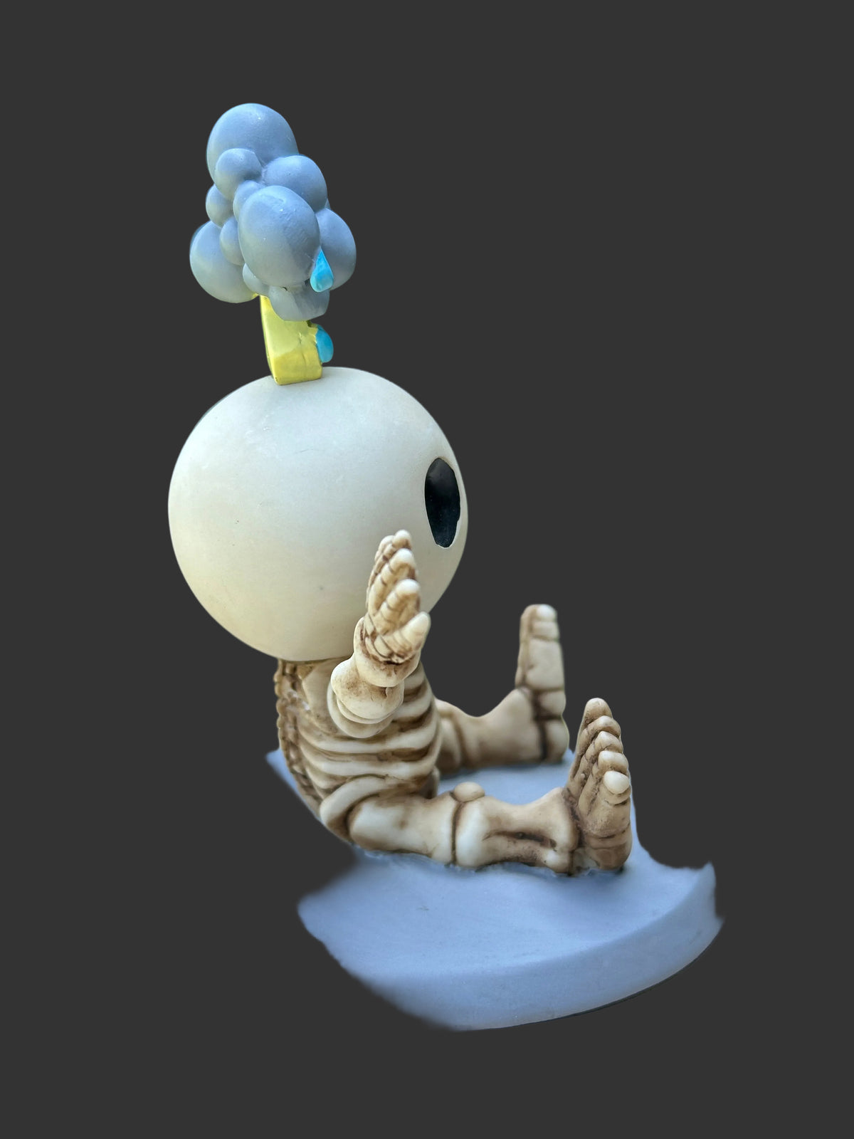 A small figurine of a seated skeleton-like character with a large round white head, pale beige limbs and face details, a light blue cloud-like top, and a pale blue base, shown on a dark gray background.