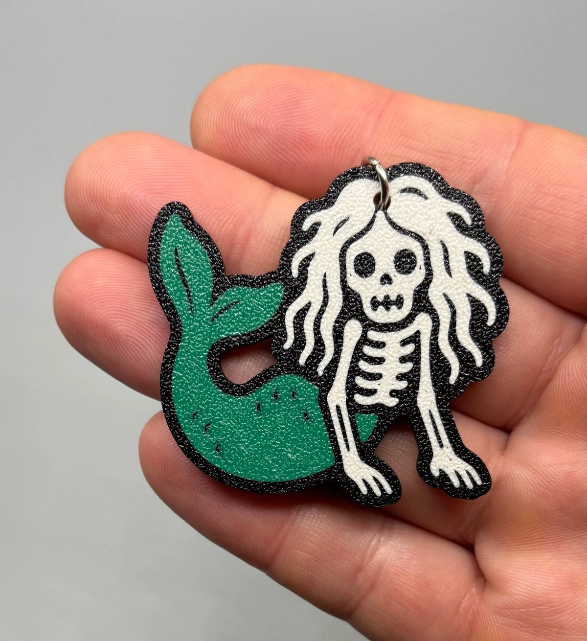 A hand holding a small decorative accessory featuring a white skeleton figure with long white hair and a green mermaid tail, outlined in black.