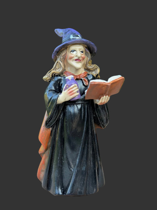 A painted miniature resin figurine of a witch in a black robe and purple hat, holding an open book, displayed on a wooden surface with a black background.