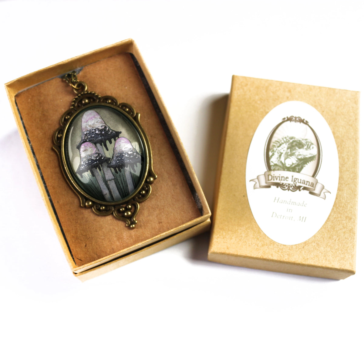 An oval pendant with an antique bronze frame displays a gothic illustration of three mushrooms glowing in a misty forest. The pendant is shown inside its brown presentation box next to its gold-colored packaging box, which features a white oval label reading 'Divine Journal Handmade in Portland, ME'.