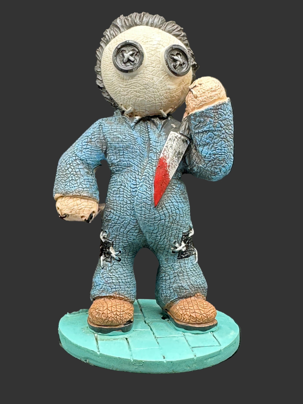 A creepy, stitched horror figurine resembling a scarecrow in a blue jumpsuit with a red-and-white striped tie, holding a knife to its face, standing on a teal circular base against a black background.