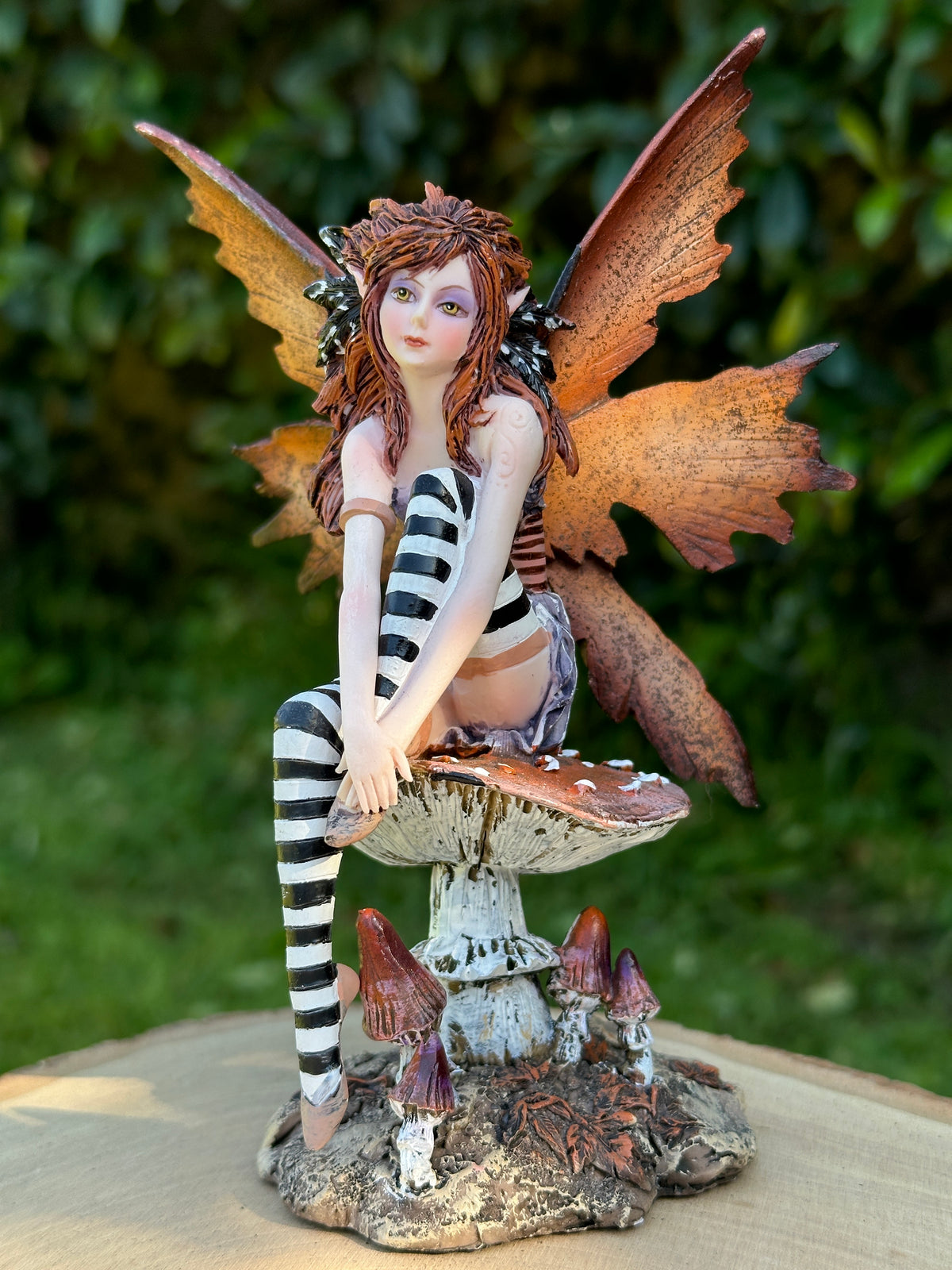 An Amy Brown fantasy fairy figurine with brown wings and striped black-and-white clothing, seated on a mushroom base with small mushrooms around it on blurred background.