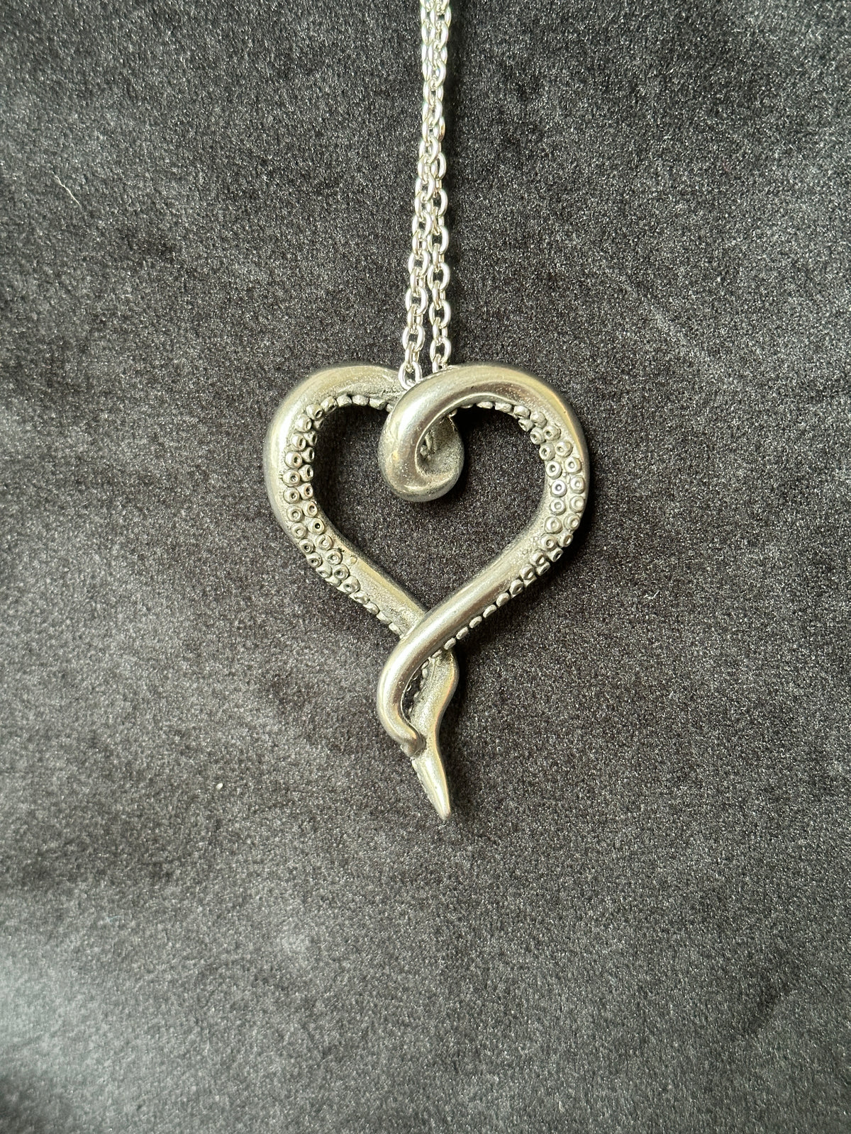 A silver Octopus pendant necklace featuring a heart-shaped design with one arm detailed in small raised dots, resting on a textured dark gray background.