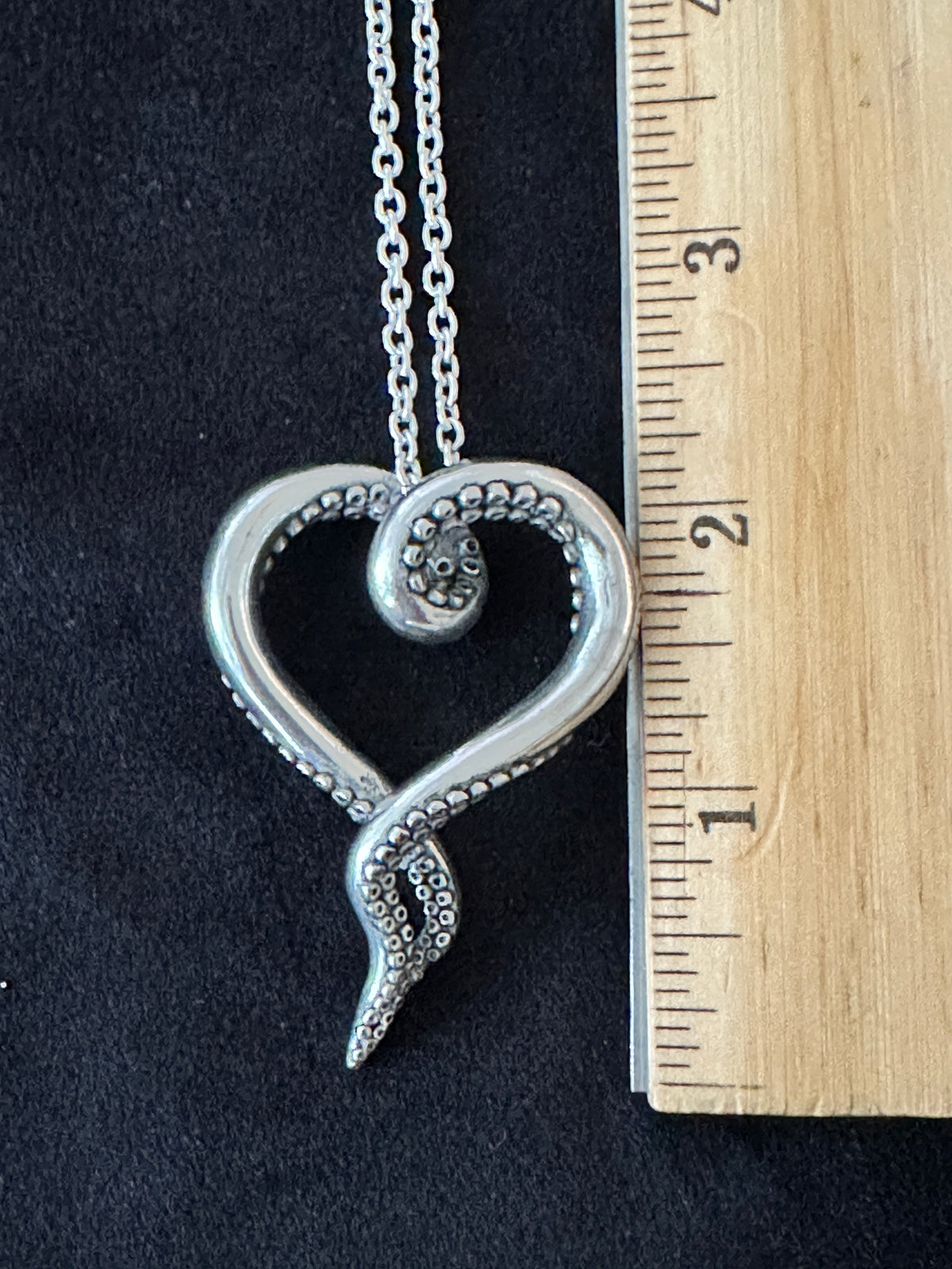 A silver heart-shaped pendant necklace with a spiral heart design, shown next to a wooden ruler on a black background to indicate size.