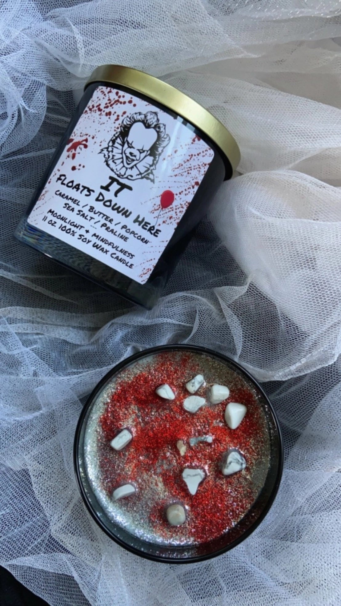 Horror themed Pennywise Candle with a label featuring a clown character and text, placed on a sheer white fabric background.