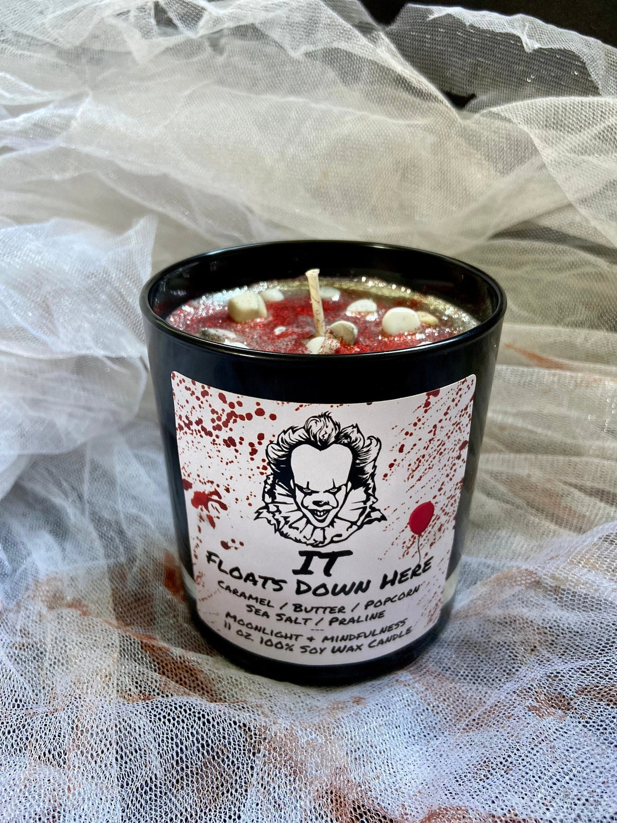 Red and White Candle with a label featuring a Pennywise character design, surrounded by sheer fabric.
