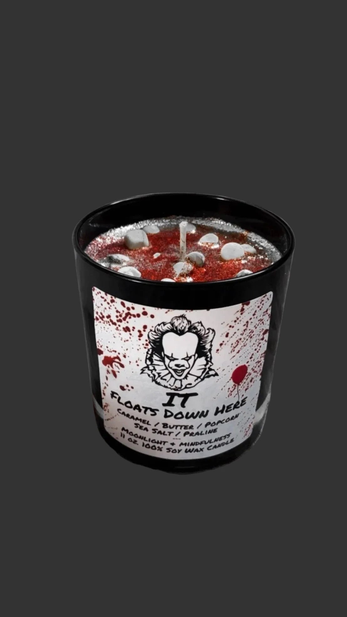 A black tin jar candle with a white label reading 'IT FLOATS DOWN HERE' and Pennywise-style artwork, filled with red wax and small white stones, set against white tulle fabric.