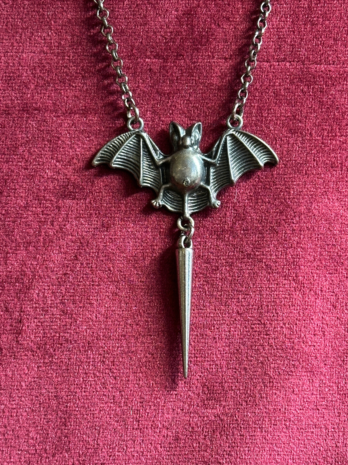 A pewter-toned bat pendant necklace with spread wings and a dangling spike charm, displayed on a textured magenta-pink fabric background.