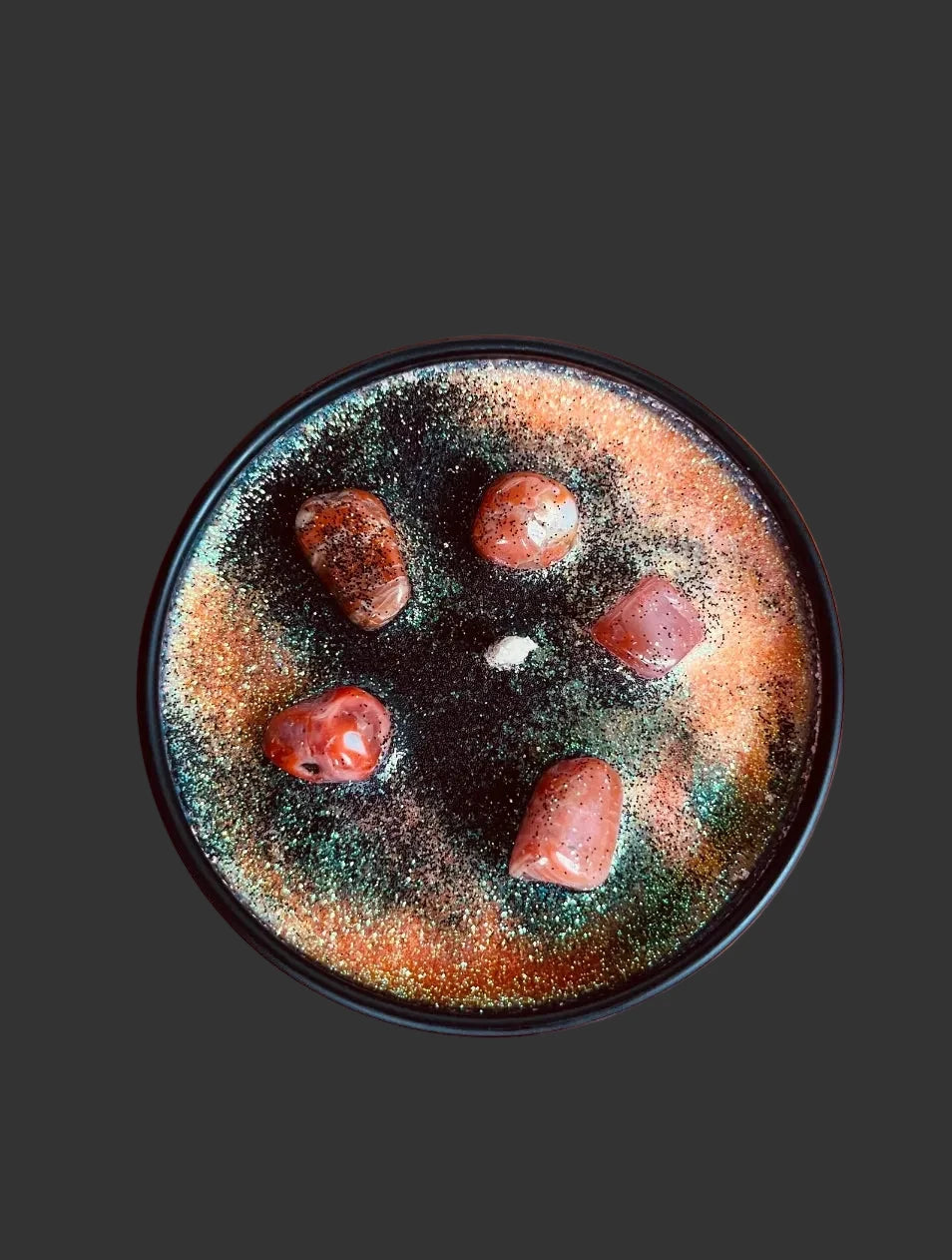 A top-down view of a round black candle with a colorful, textured wax surface featuring orange, green, and black tones, decorated with five reddish-brown stones, set against a warm orange background with a pumpkin partially visible in the corner.