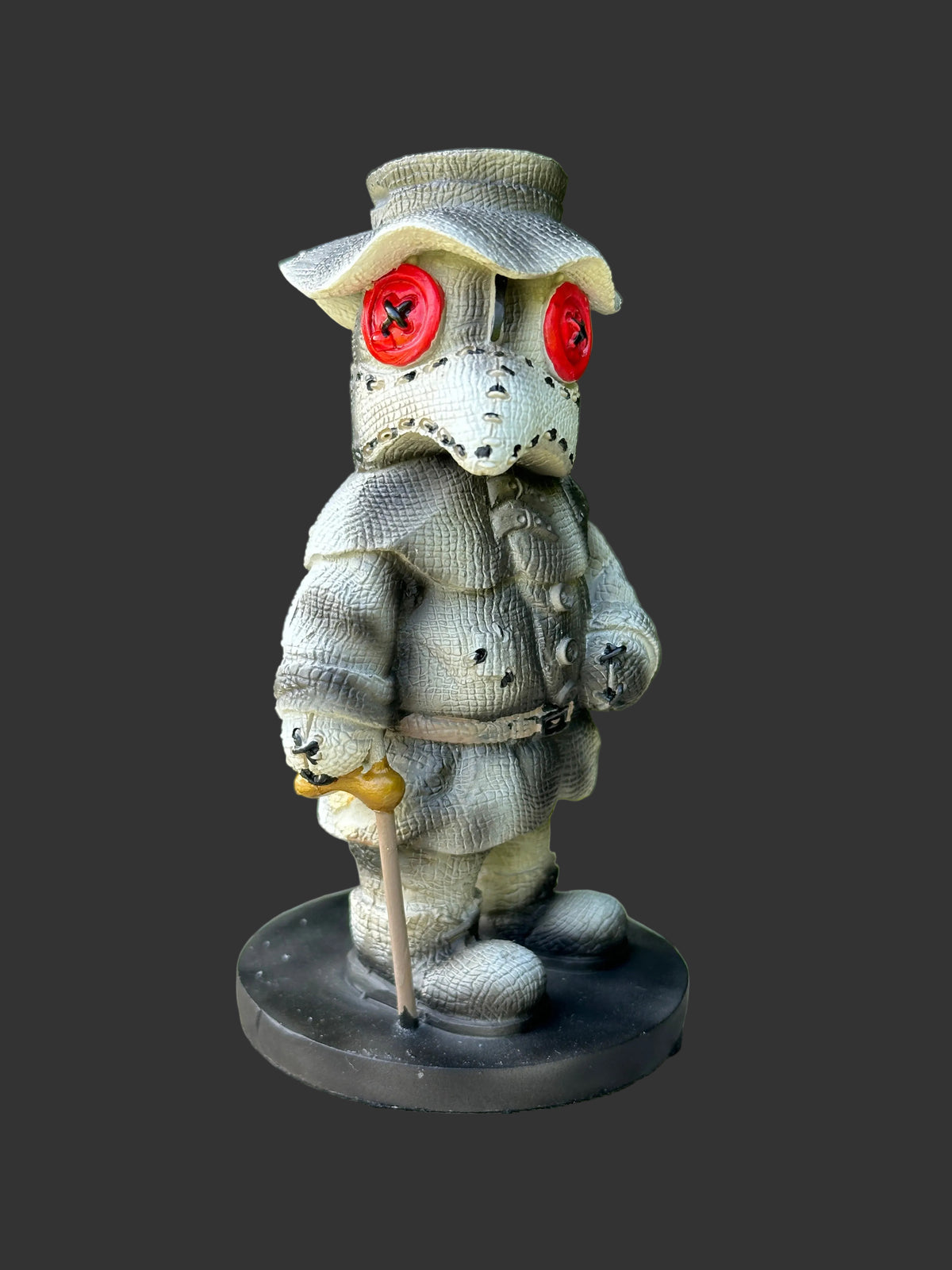 A small decorative figurine of a stitched bird-like character wearing a hat and coat, standing on a round black base and holding a cane, photographed outdoors on a wooden surface with blurred green foliage behind it.