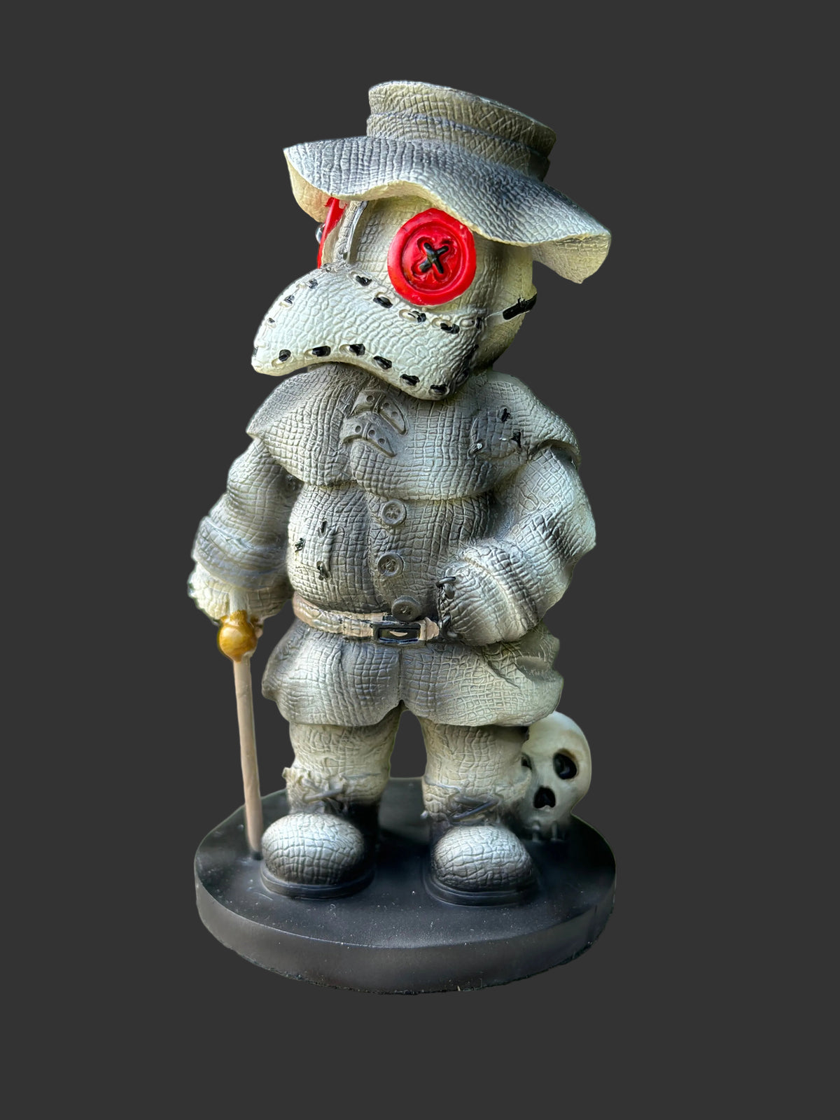 A stitched scarecrow figurine wearing a wide-brimmed hat and a red circular eye patch, standing on a black base with a cane and a small skull at its side, photographed outdoors on a wooden surface with blurred green foliage in the background.