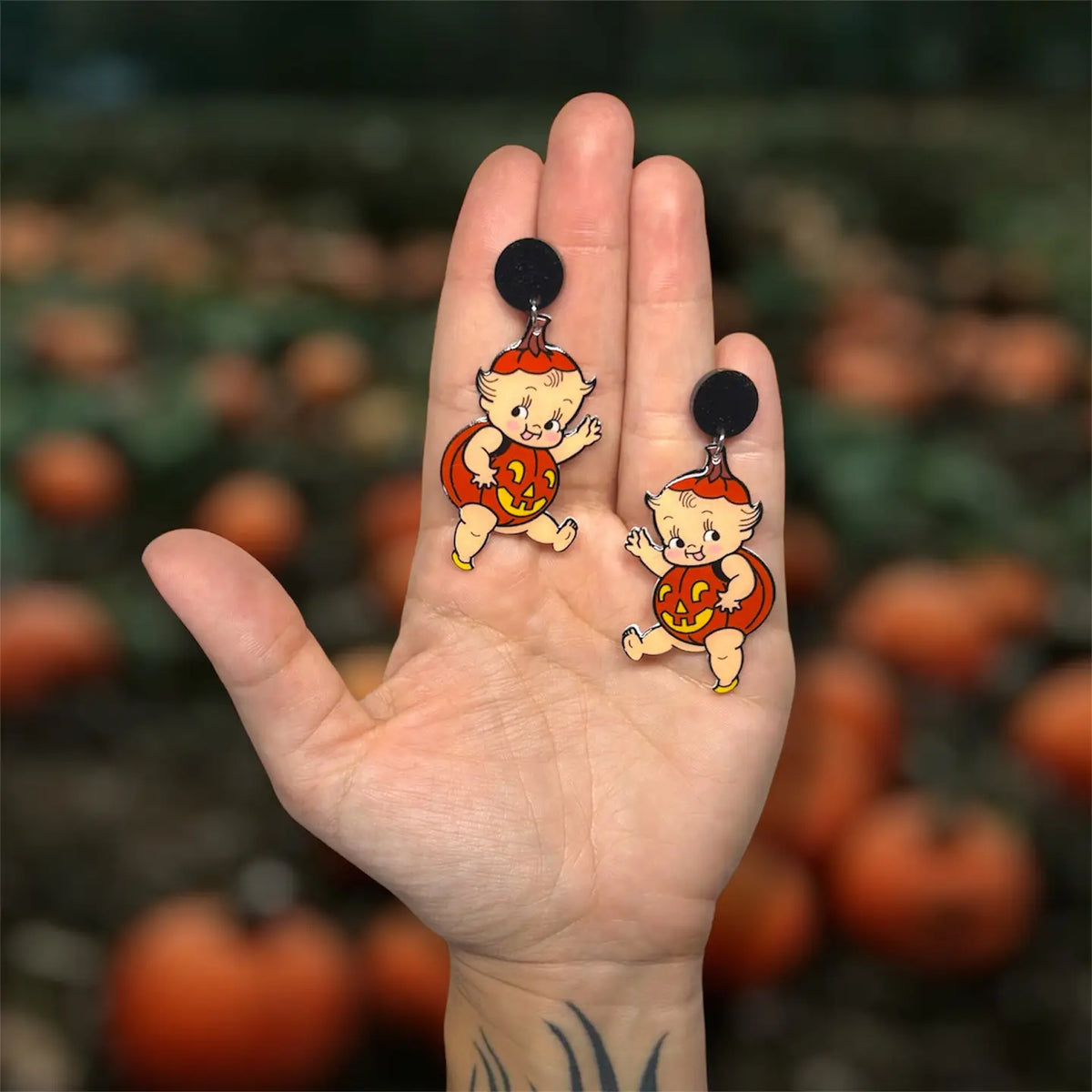 A pair of novelty dangle earrings featuring Kewpie cartoon baby pumpkins wearing orange and yellow outfits with the number 12, held in a person's hand against a Black pumpkin patch background