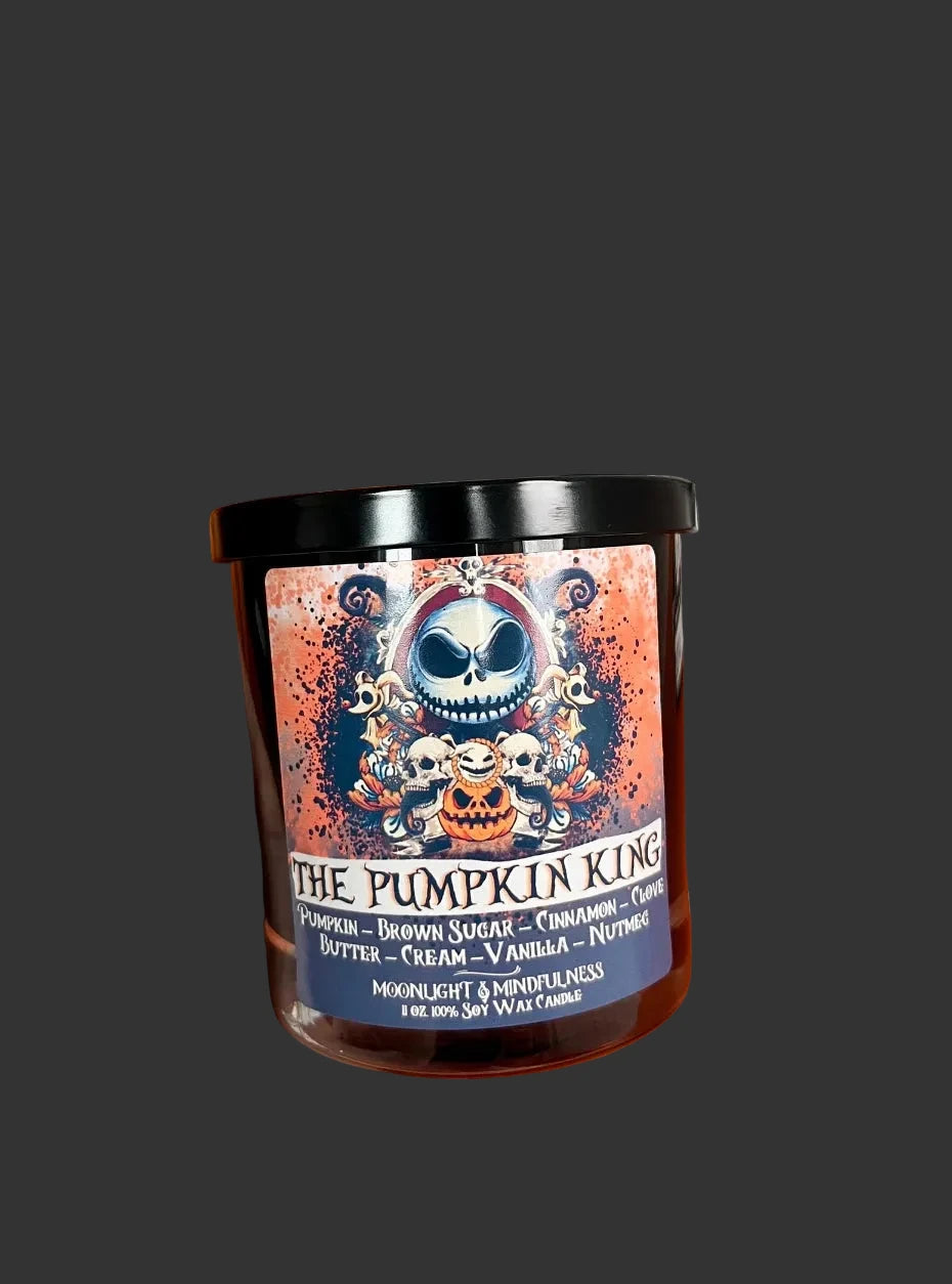 A hand holds a candle jar labeled 'The Pumpkin King' against a Halloween-themed background with a pumpkin, brick wall, spider clock, and fall decorations.