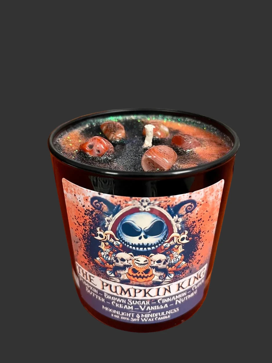 A Halloween-themed jar candle labeled 'The Pumpkin King' sits on an orange fabric surface with decorative pumpkins and a spiderweb clock in the background. The candle has a black glass container, a colorful label with a skull illustration, and visible dried spices on top.