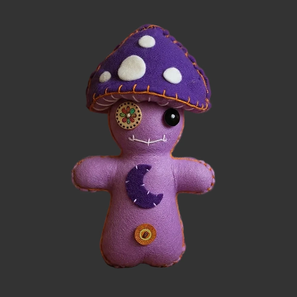 A purple felt mushroom-shaped poppet doll with white spots, a crescent moon on its chest, and a gold button, posed on a patterned rug with candles and bohemian decor in the background.