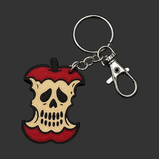 A skull-shaped keychain charm designed to look like a bitten red apple, featuring a beige skull in the center with black and red details, attached to a silver metal key ring and lobster clasp, displayed on a dark textured stone background.