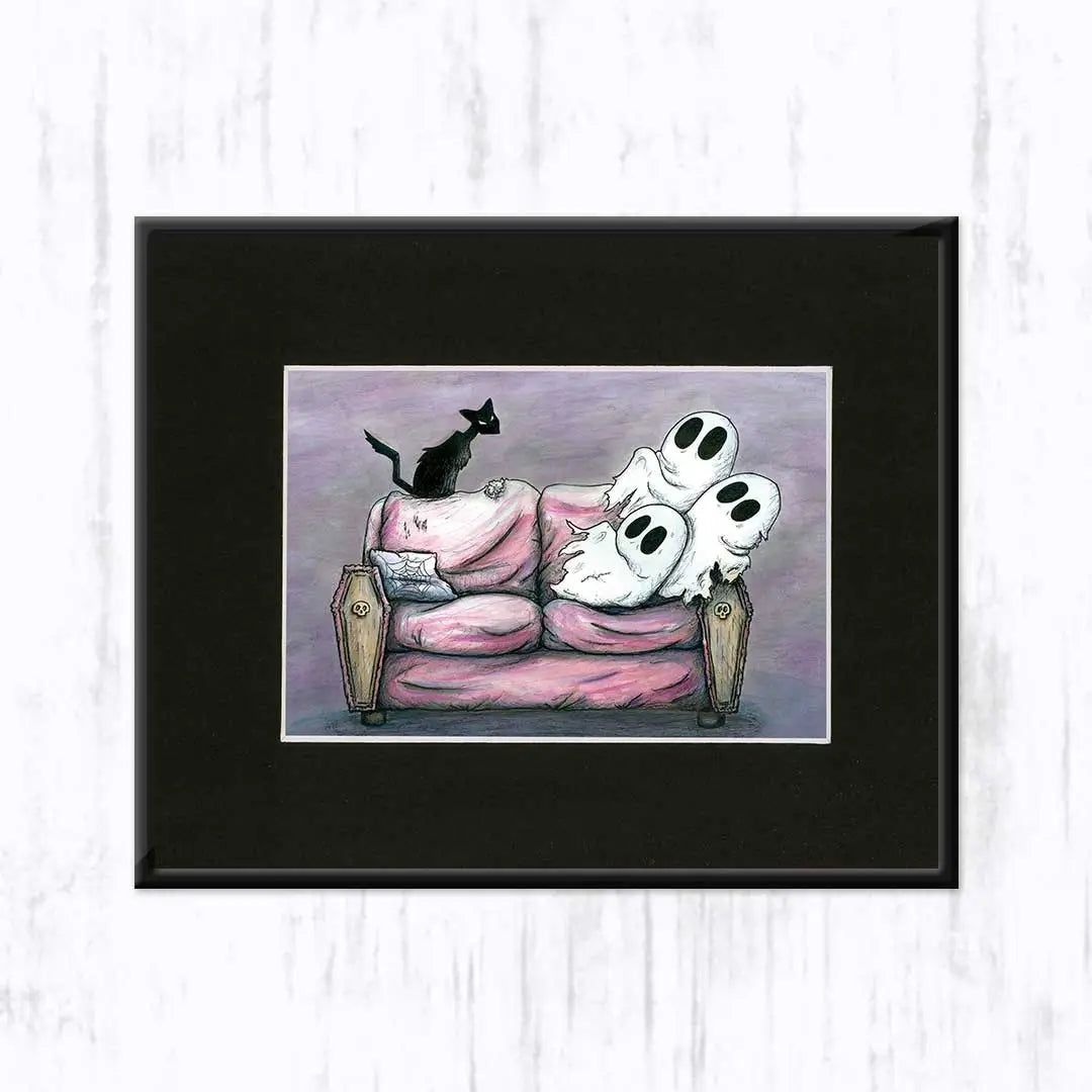 A framed art print featuring a whimsical illustration of three white ghosts resting on a pink coffin-shaped couch, with a black cat perched on the back, set against a light gray wood-textured background.