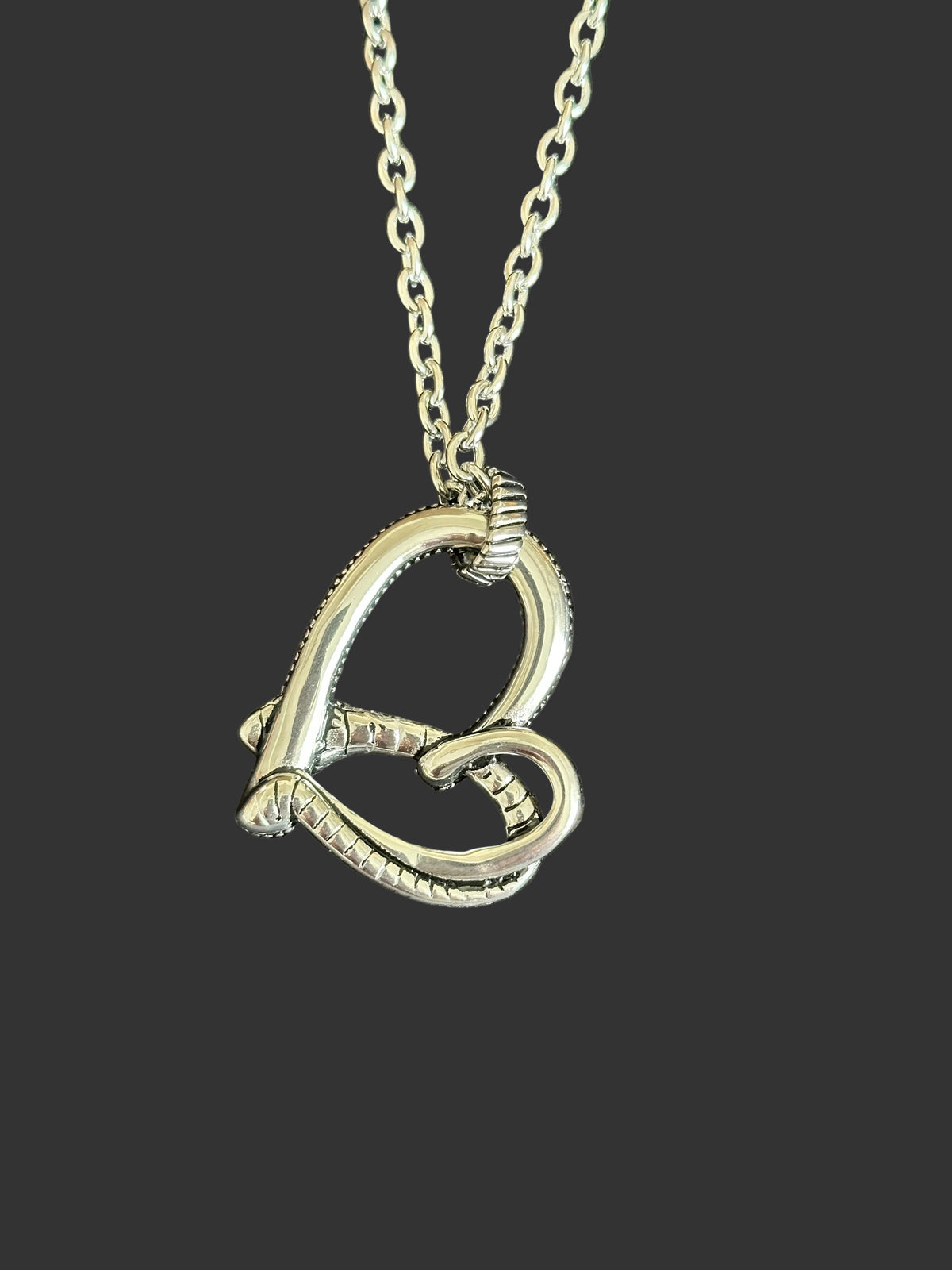 A silver-toned metal pendant necklace featuring two interlocking heart shapes on a matching chain, displayed on a dark textured fabric background.