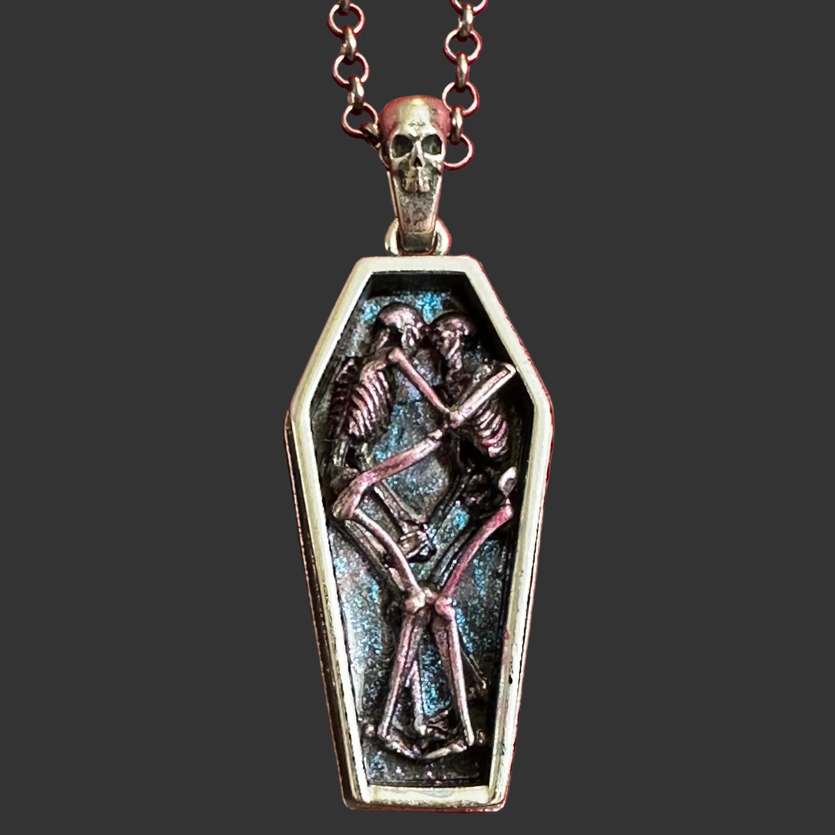 A gothic coffin pendant necklace featuring two interlocking purple skeleton figures on a silver chain featuring a silver skull displayed on a textured red velvet background.