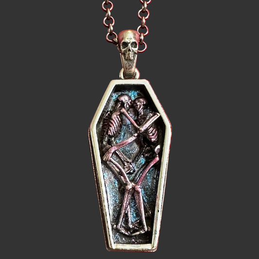 A gothic coffin pendant necklace featuring two interlocking purple skeleton figures on a silver chain featuring a silver skull displayed on a textured red velvet background.