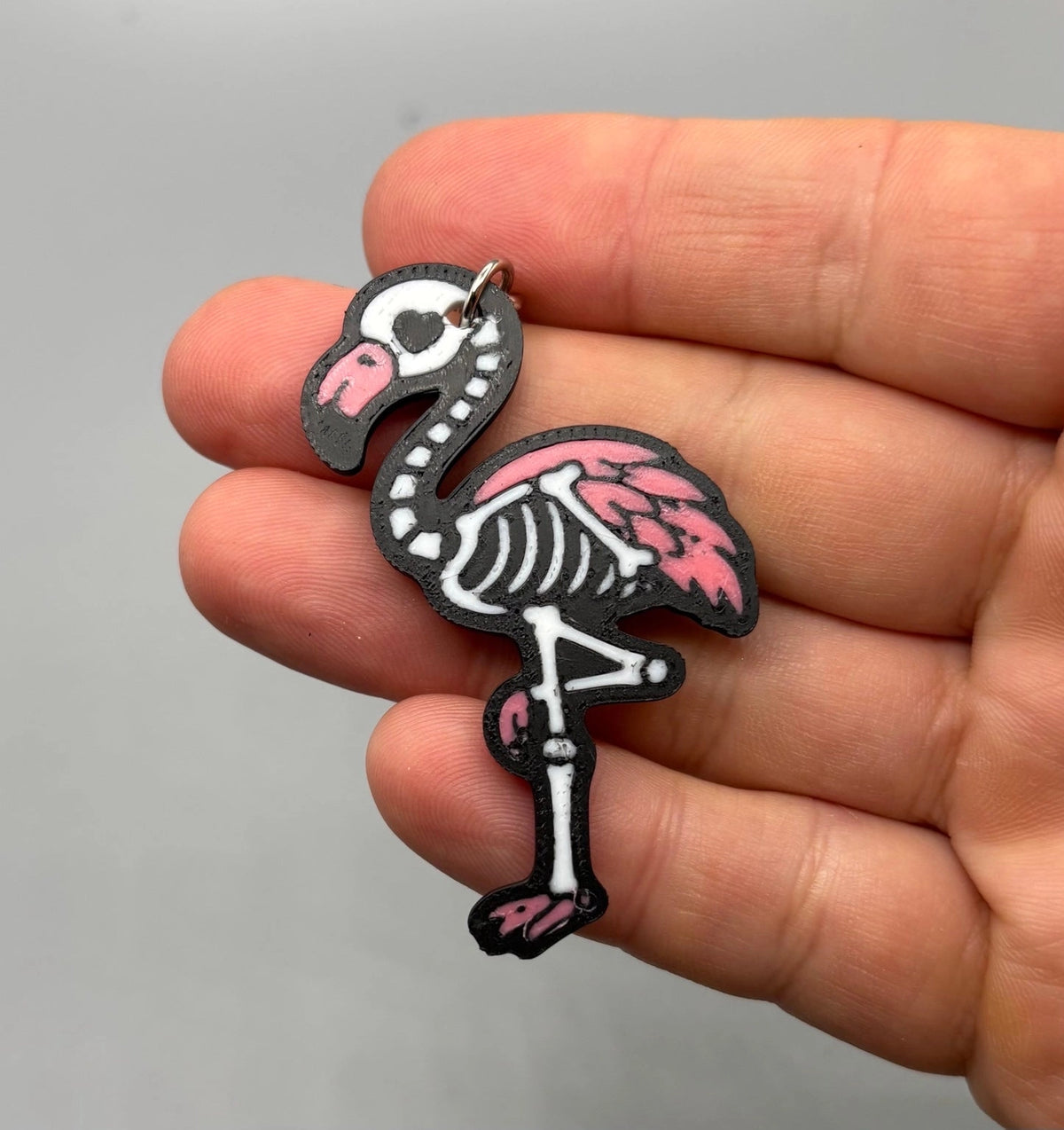 A hand holding a small flamingo-shaped charm or pendant with a skeleton design, featuring a black outline, white bones, and pink feathers, against a plain gray background.