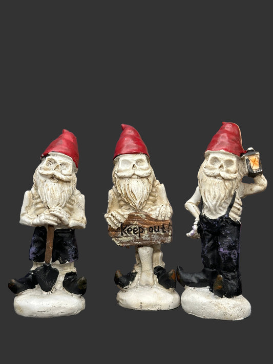 Three Halloween garden gnome miniature figurines with skull faces and red hats, standing on a dark surface against a black backdrop. The left gnome holds a shovel, the center holds a 'Keep out' sign, and the right holds a lantern.