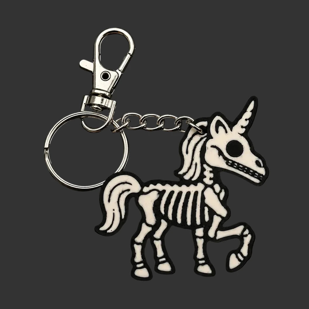A skeleton unicorn keychain  or charm with a silver metal clasp and key ring, featuring a white and black unicorn charm with skeletal details, displayed on a dark textured slate background.