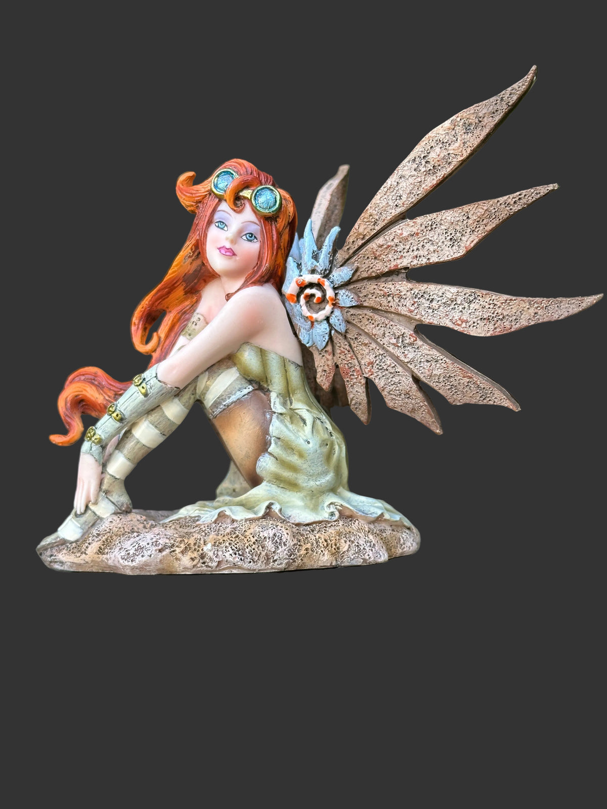 A fantasy Amy Brown steampunk fairy figurine with orange hair, large brown wings, and a striped dress, posed on a stone-like base outdoors against a blurred green foliage background.